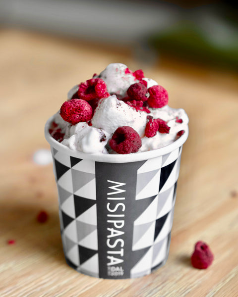 Ice cream with raspberries in a cup labeled 'Misipasta' on a wooden surface.